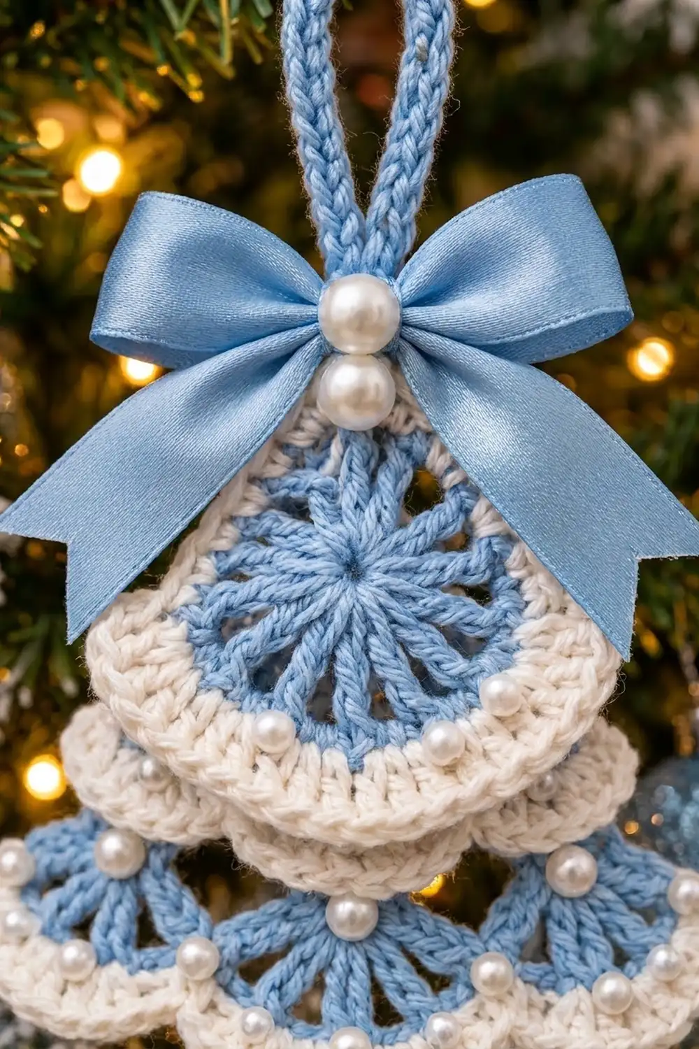 Handmade crochet Christmas ornament in cream and blue with pearl-adorned bow, hanging against warm glowing holiday lights.
