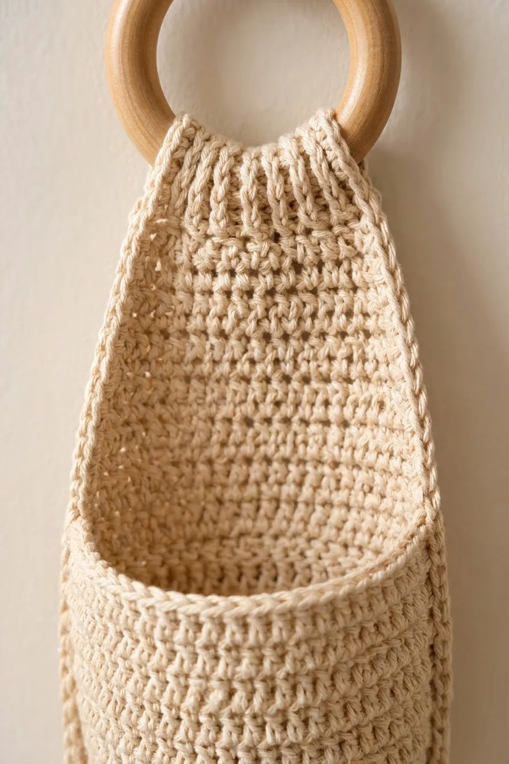 Crocheted cream-colored bag with detailed texture hangs from a natural wooden ring handle on a neutral backdrop.