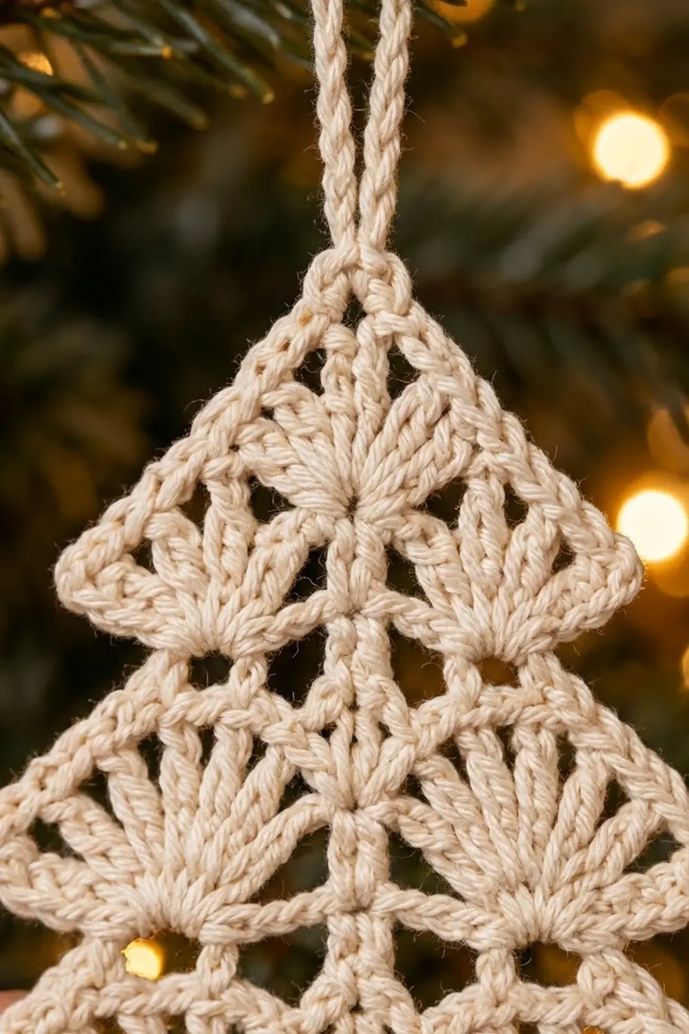 Cream-colored crocheted tree ornament with intricate knotted pattern, hanging against blurred evergreen and warm golden lights.