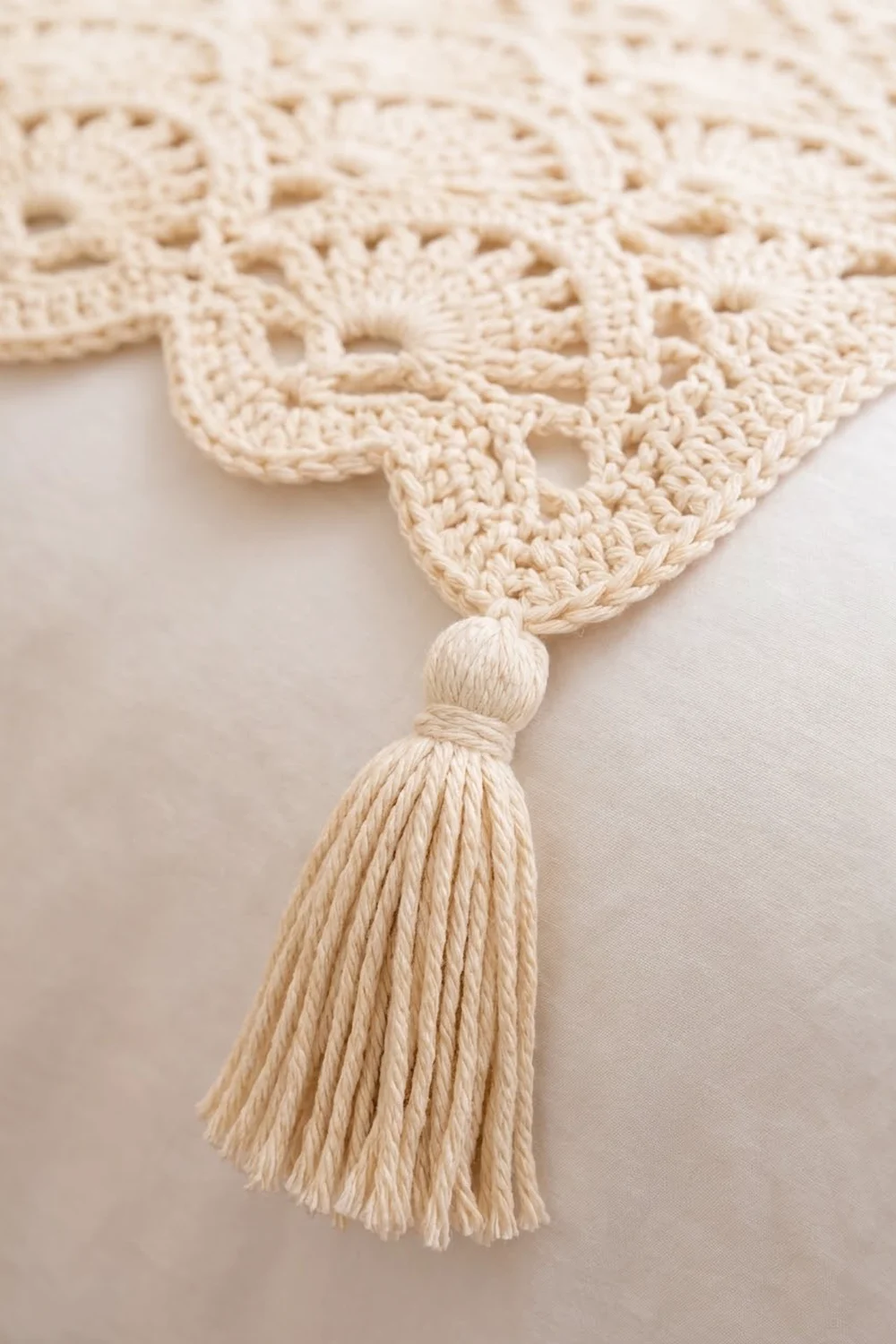 Cream-colored crochet detail with delicate openwork pattern and a knotted tassel against a soft neutral background.