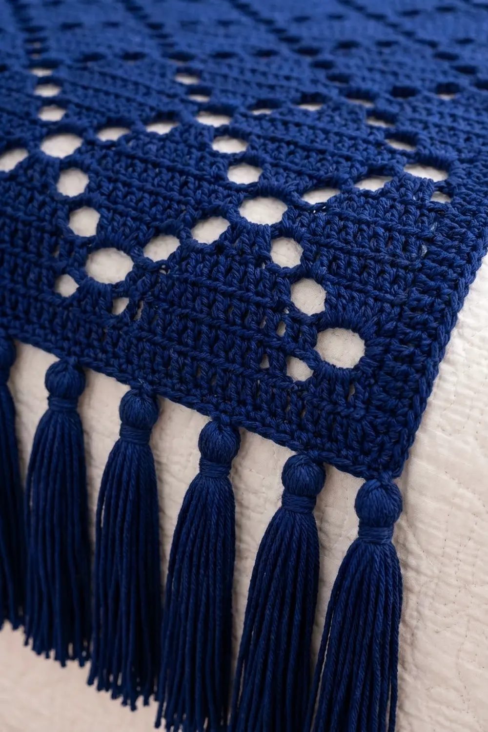 Close-up of navy blue crocheted lace with circular eyelet holes and matching yarn tassels on white background.