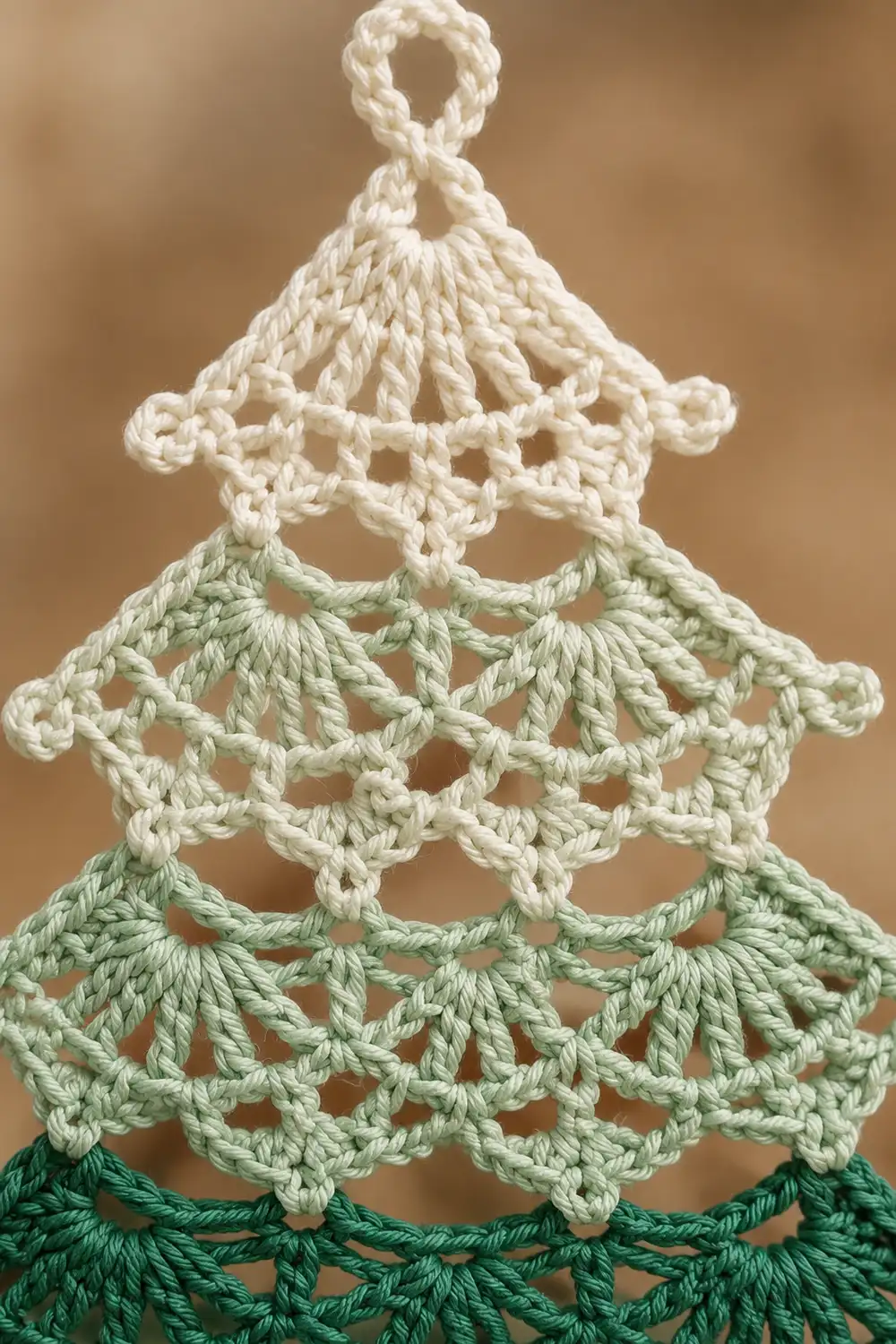 Crocheted Christmas tree ornament with cream, light green, and dark green layered scalloped sections against a blurred tan background.