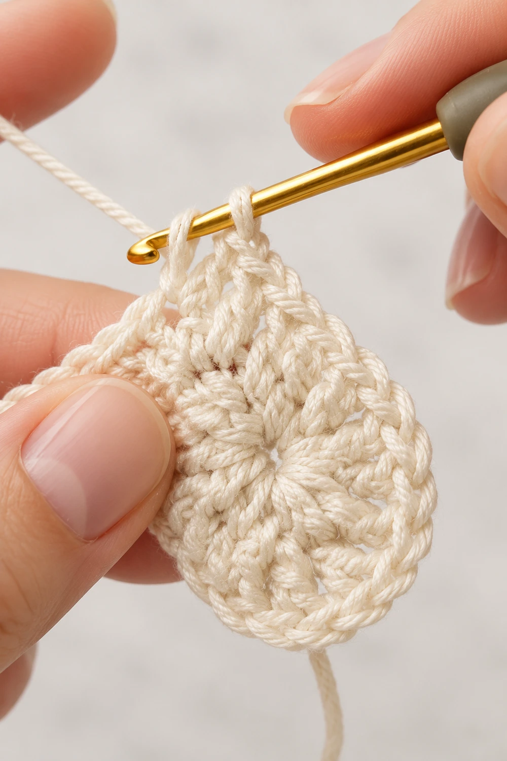 Close-up of cream-colored crocheted swatch on gold crochet hook held in hands against white background.
