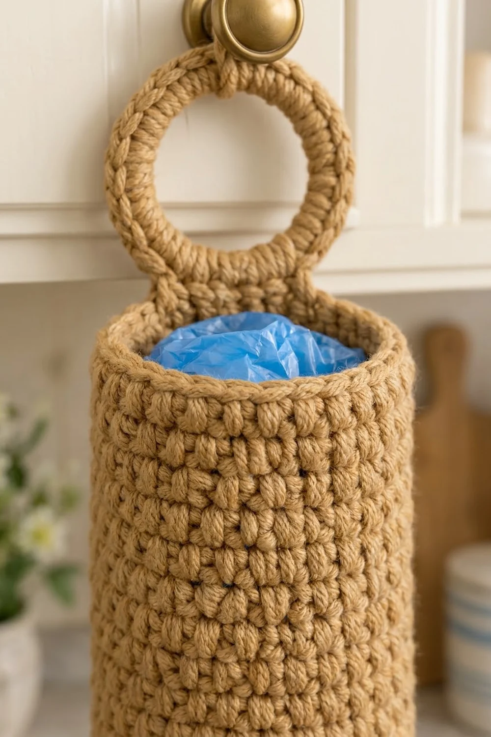 Tan crocheted hanging basket with textured stitching, brass hook attachment, and visible blue plastic lining against white paneling.