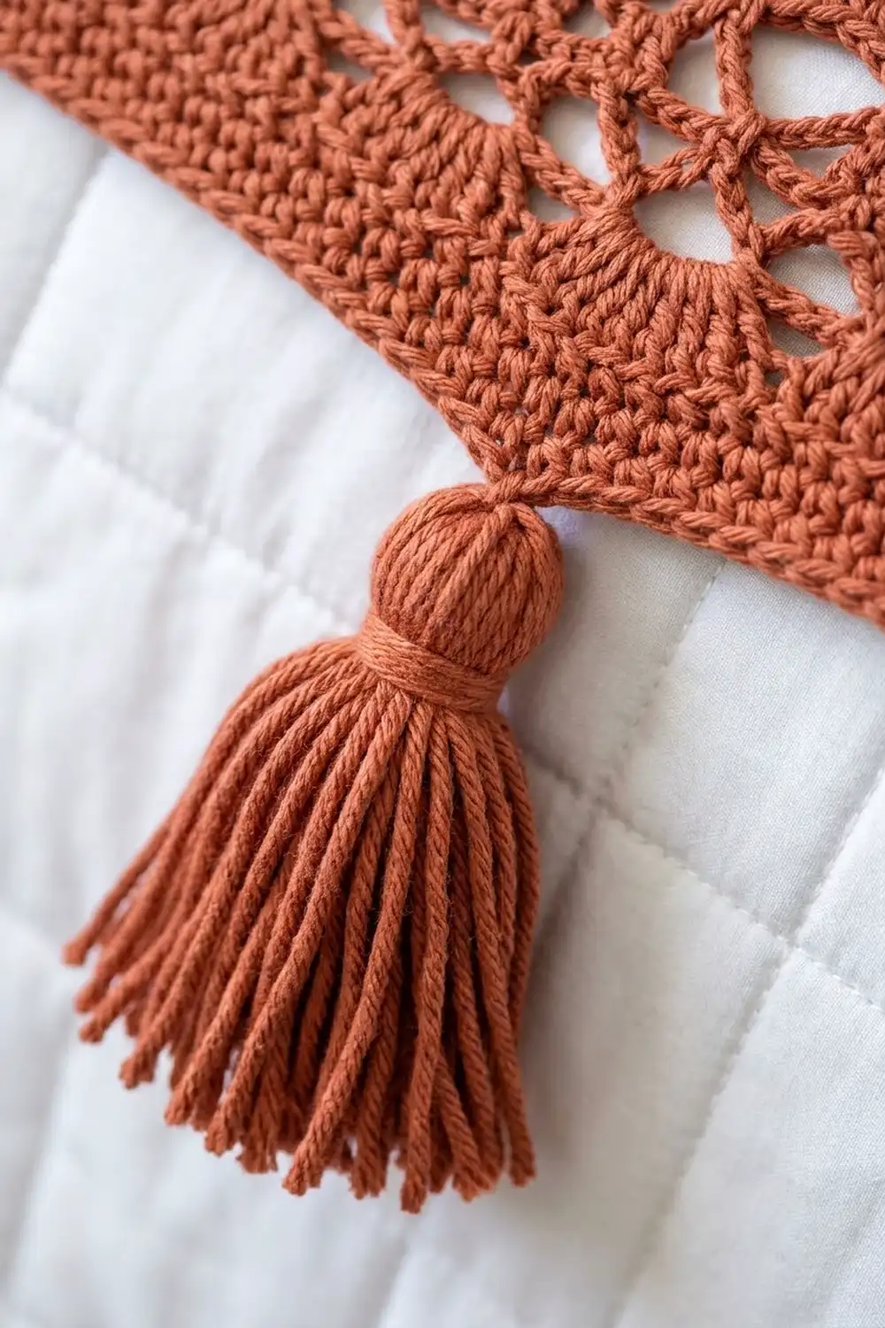Rust-colored crochet detail with delicate lacework stitches and a fringe tassel on white linen background.