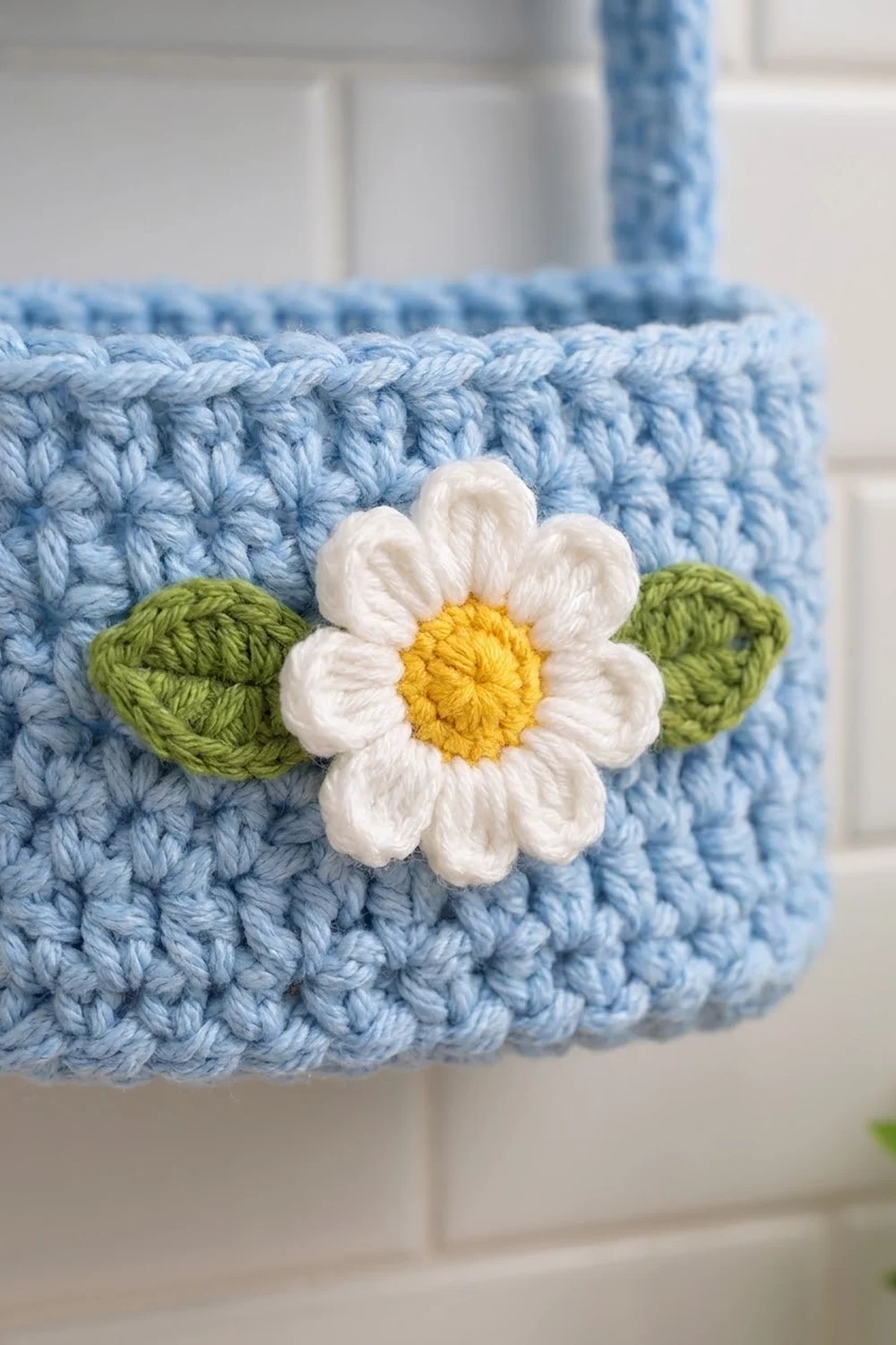 Light blue crocheted basket decorated with a white and yellow daisy flower with green leaves.