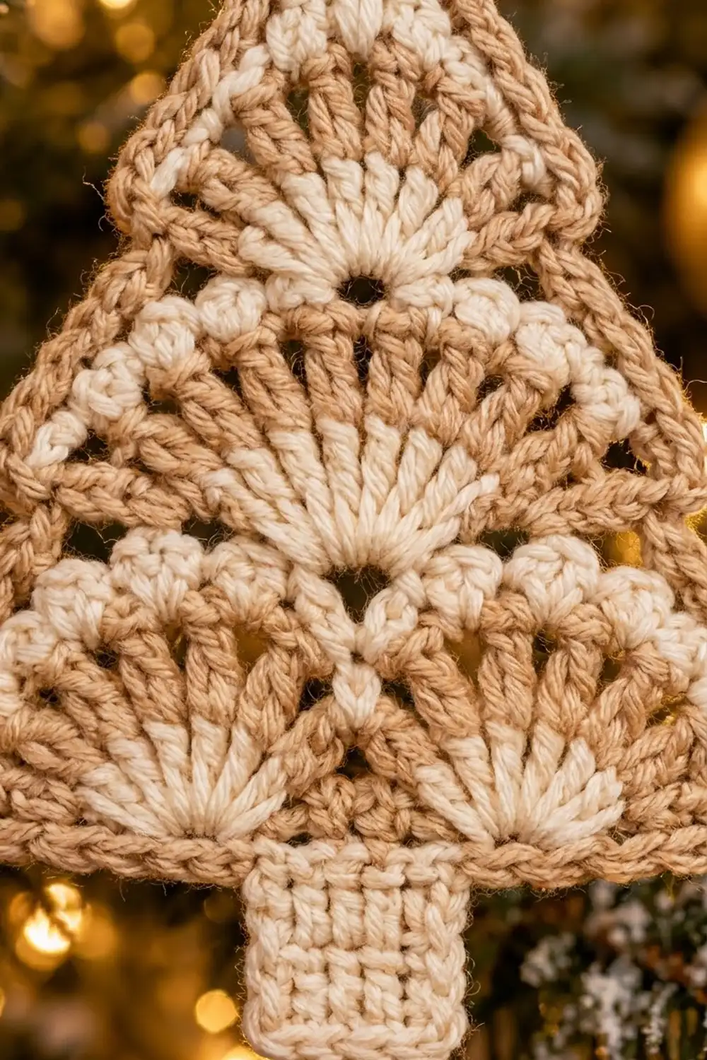 Crocheted Christmas tree ornament in cream and tan yarn with warm golden bokeh lights blurred in background.