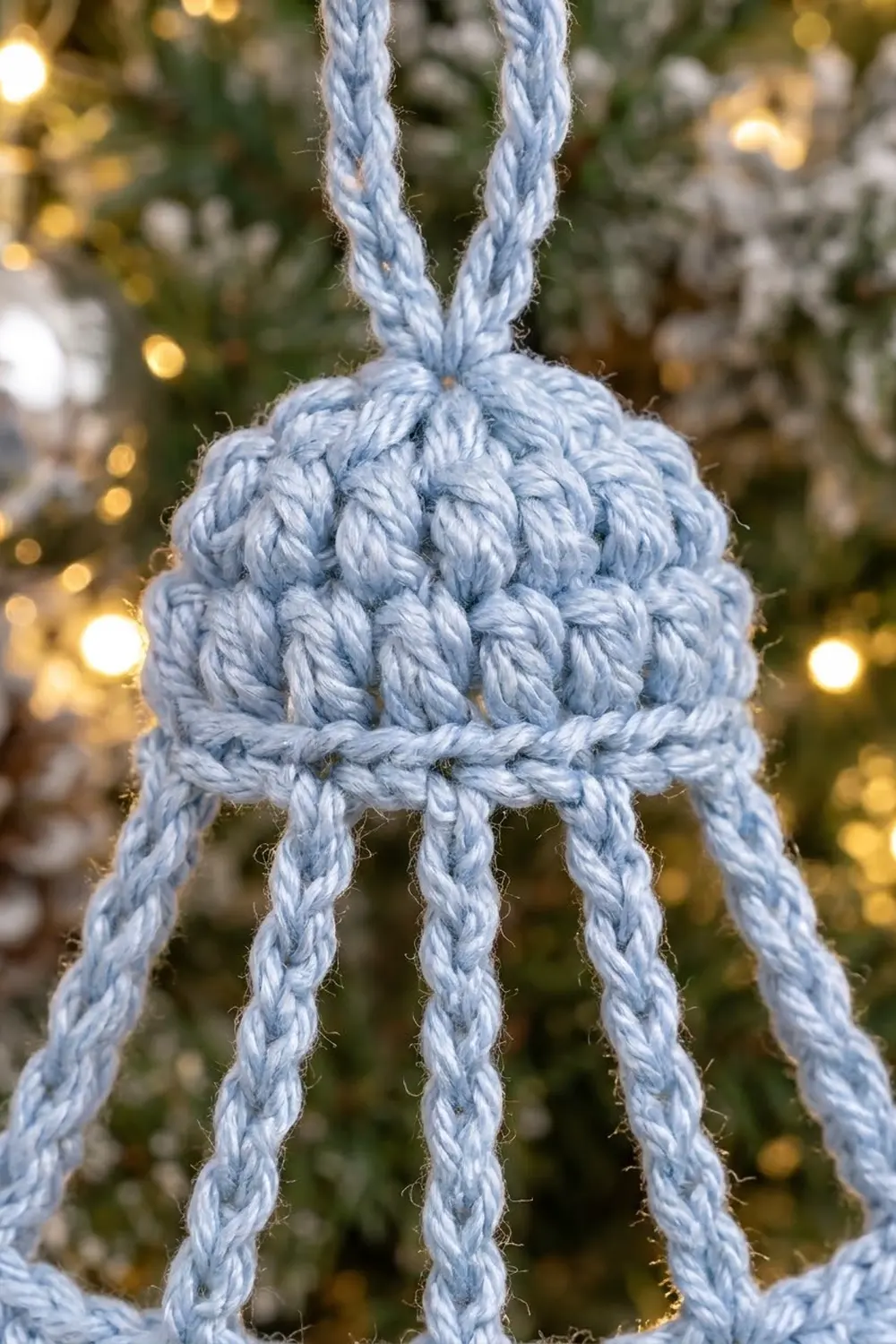 A light blue crocheted bell ornament with textured stitches and braided straps hangs before twinkling holiday lights and evergreen branches.