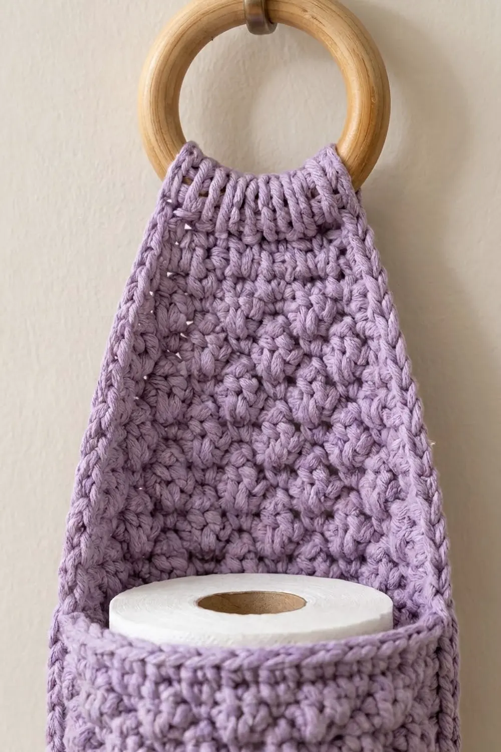 Lavender crocheted toilet paper holder with natural wood ring displaying a white tissue roll inside.