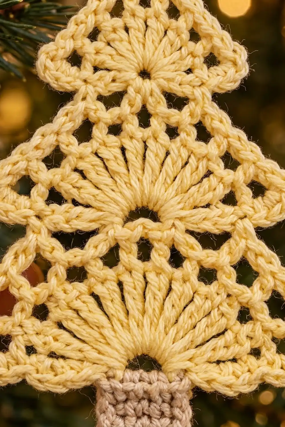 Cream-colored crocheted ornament with intricate lacy geometric pattern and beige stem, photographed against a blurred holiday background.
