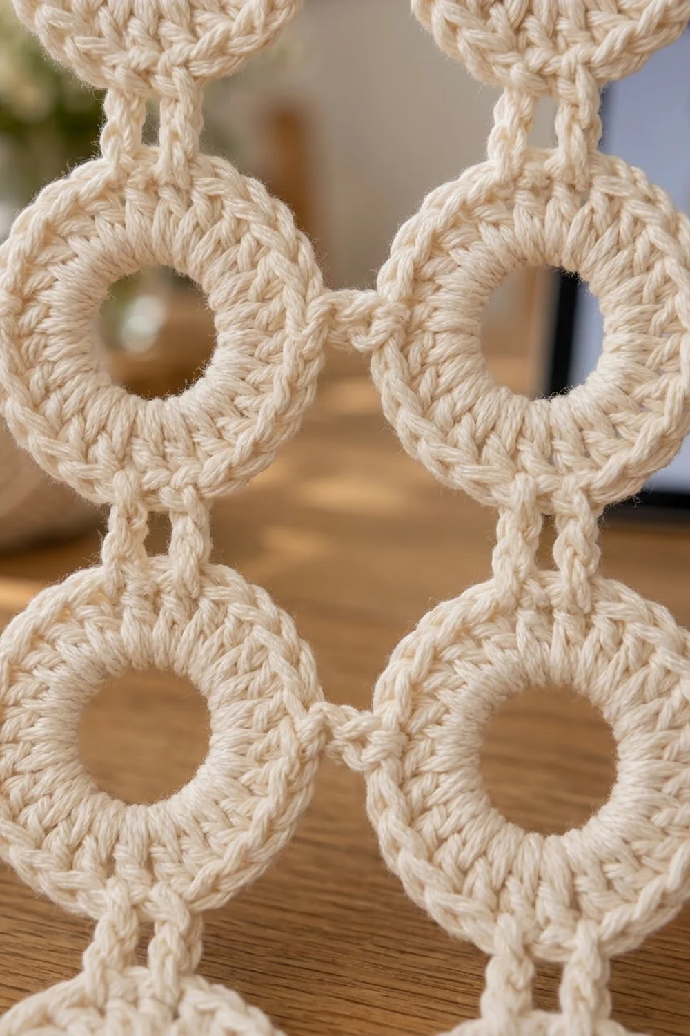 Cream-colored crocheted rope features a geometric chain pattern with circular linked rings on a warm wooden background.