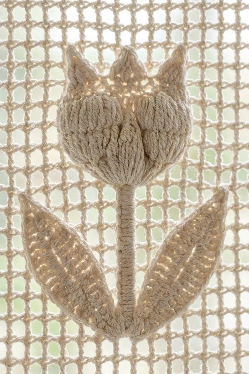 Hand-crocheted beige tulip with rope-textured petals and leaves on a light open-mesh backdrop.