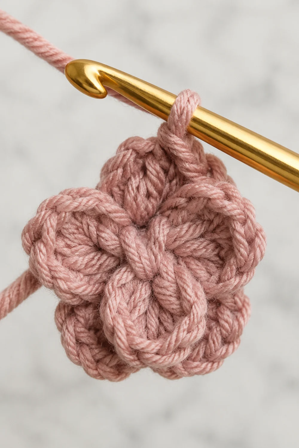 A crocheted rose ornament in dusty pink yarn hangs from a gold crochet hook against a blurred white background.