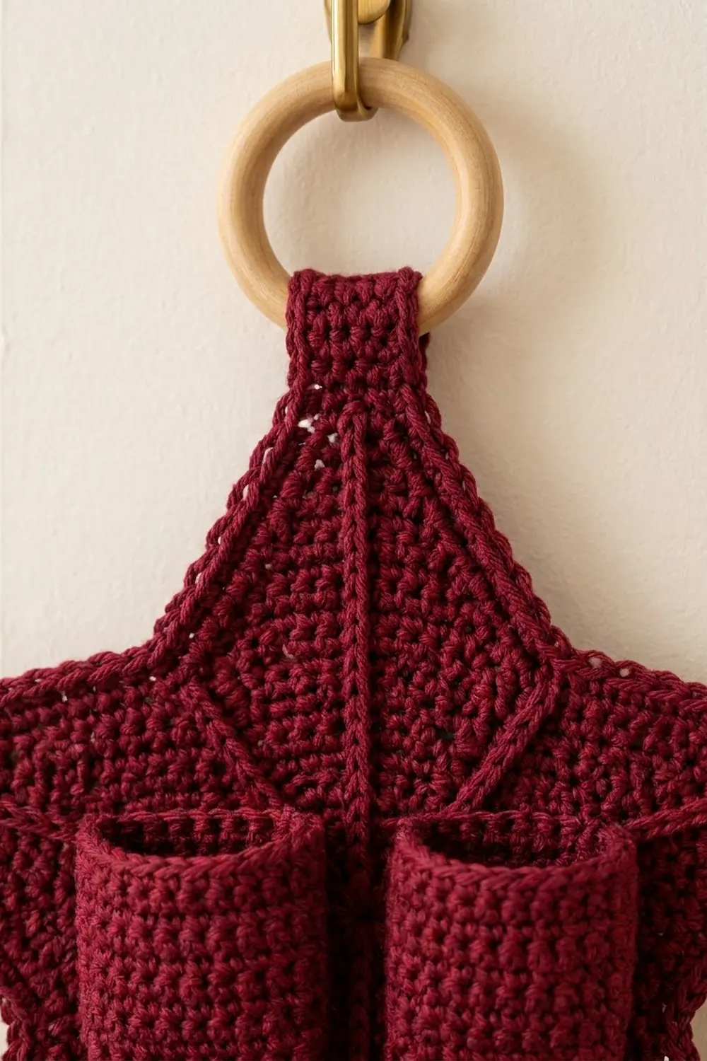 Deep burgundy crocheted fabric with open lace pattern hangs from natural wooden ring on brass hook against cream wall.