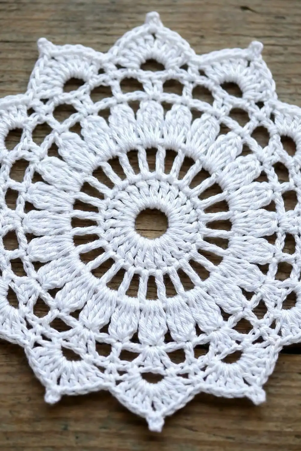 White crocheted doily with intricate openwork circular pattern and scalloped edges rests on a weathered wooden surface.