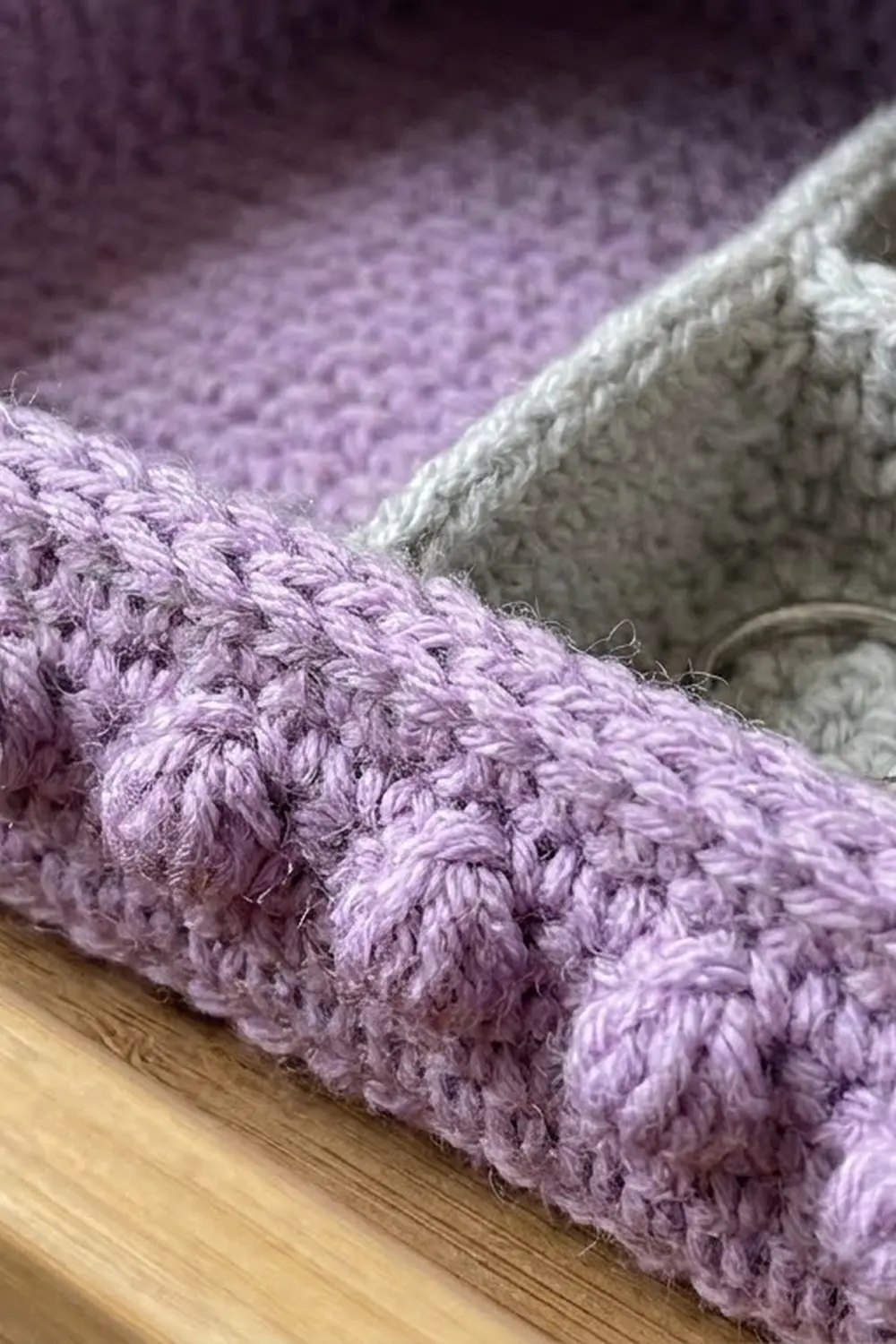 Lavender crocheted blanket with chunky textured stitches paired with cream yarn on light wood flooring.