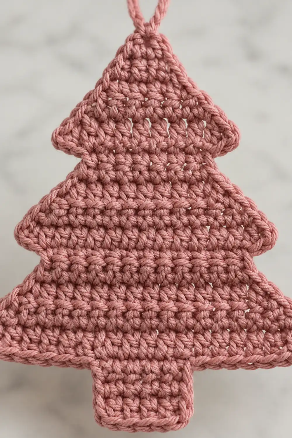 A pink crocheted Christmas tree ornament with a braided hanging loop displayed against a blurred white background.