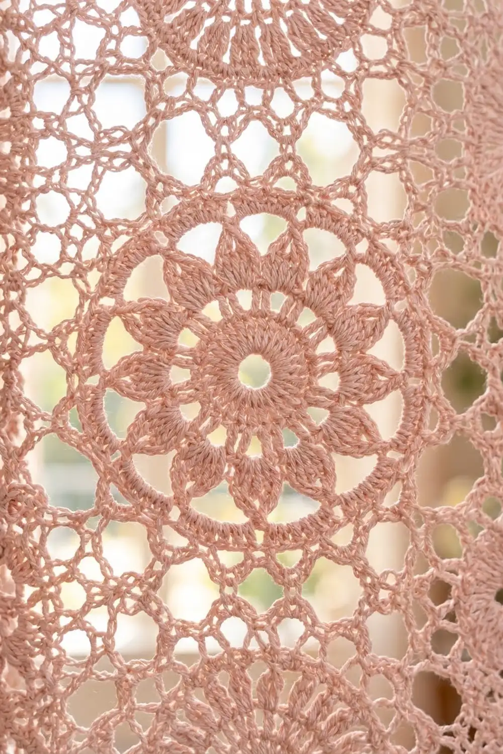 Dusty rose crocheted lace with a central flower motif and geometric openwork, showing soft bokeh background through the spaces.