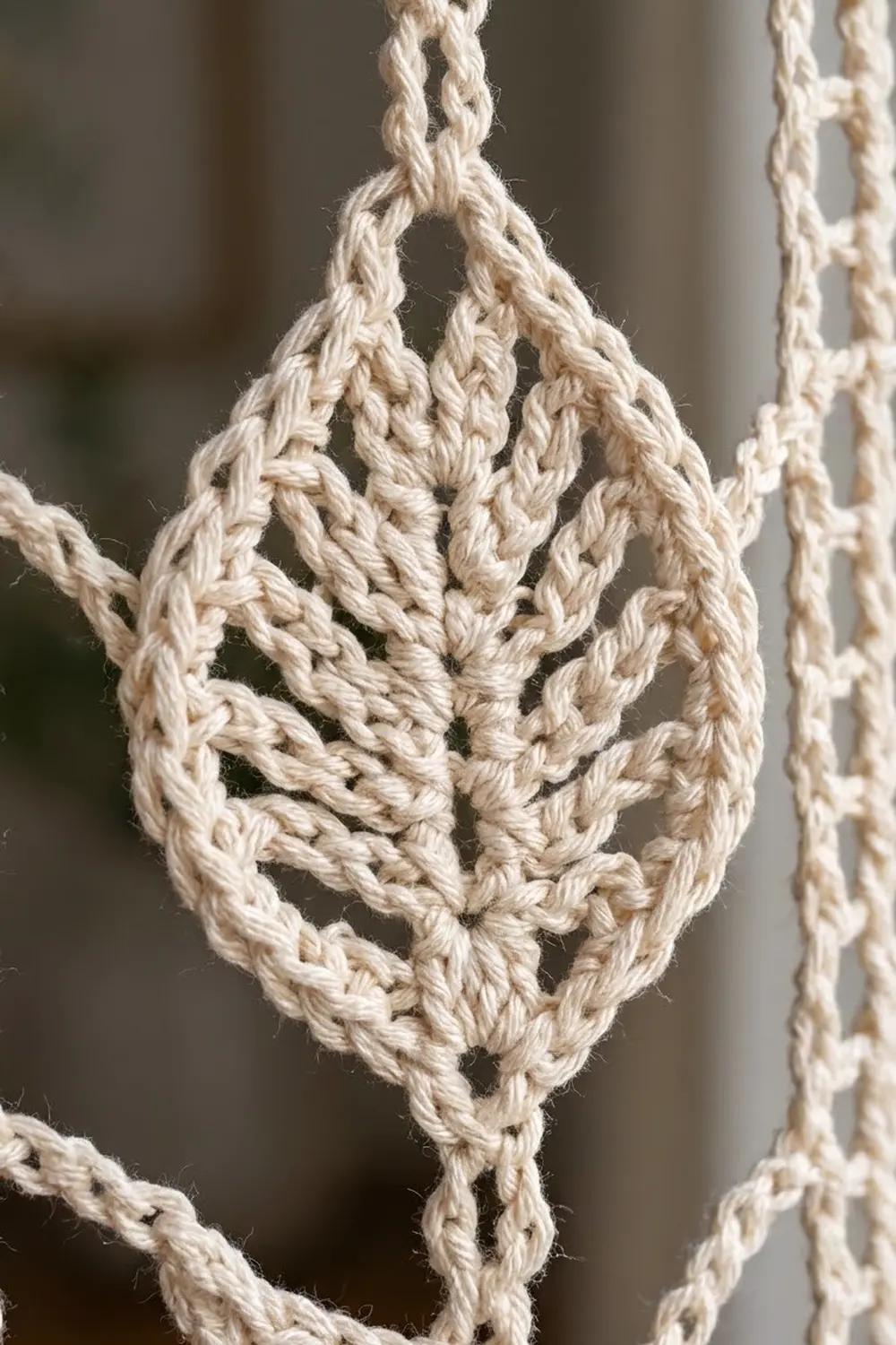 Close-up of cream-colored crocheted bag displaying detailed braided stitchwork, hanging from twisted rope against a soft neutral background.