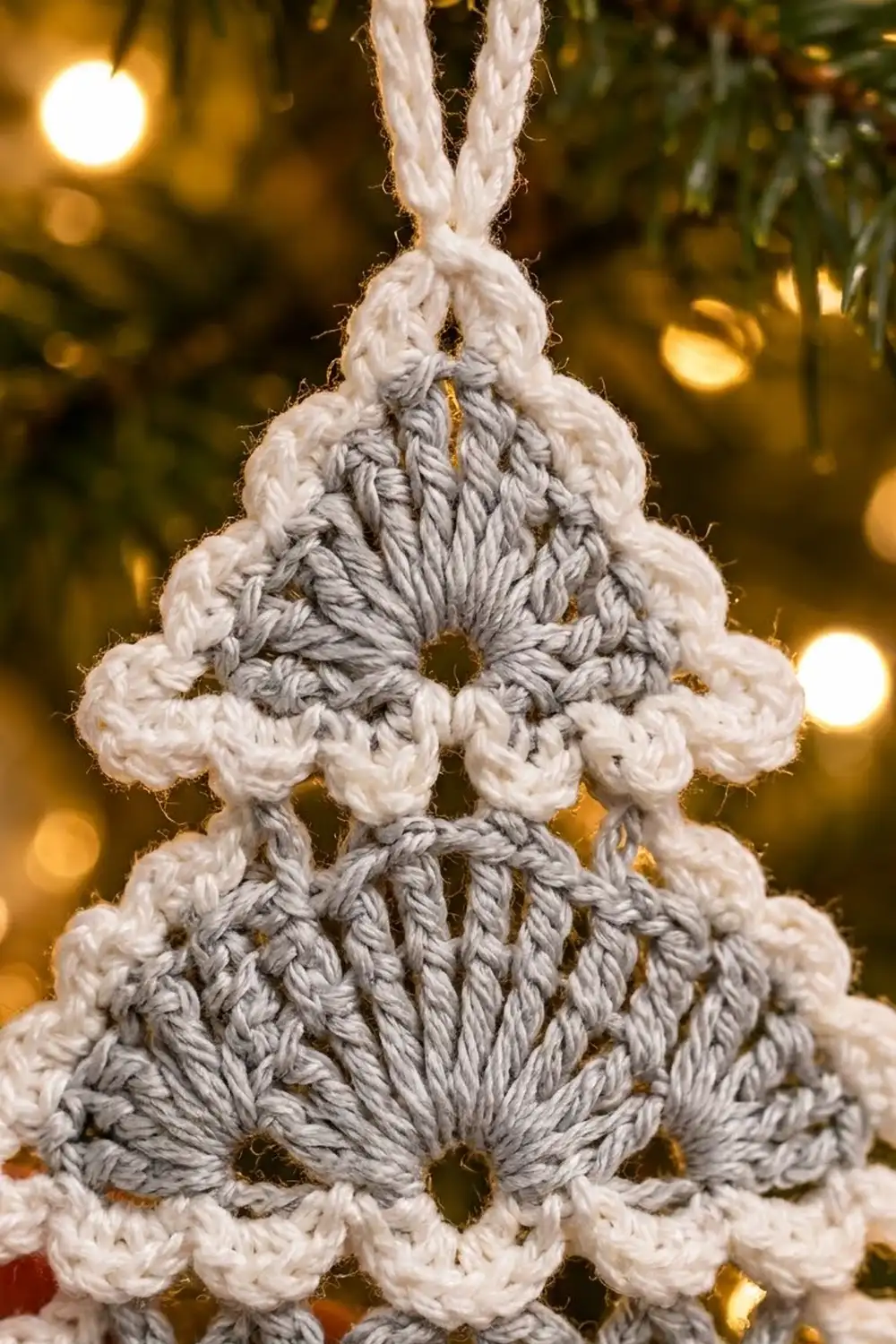 Handmade crocheted Christmas tree ornament in cream and gray yarn with braided details, hung against warm golden bokeh holiday lights.