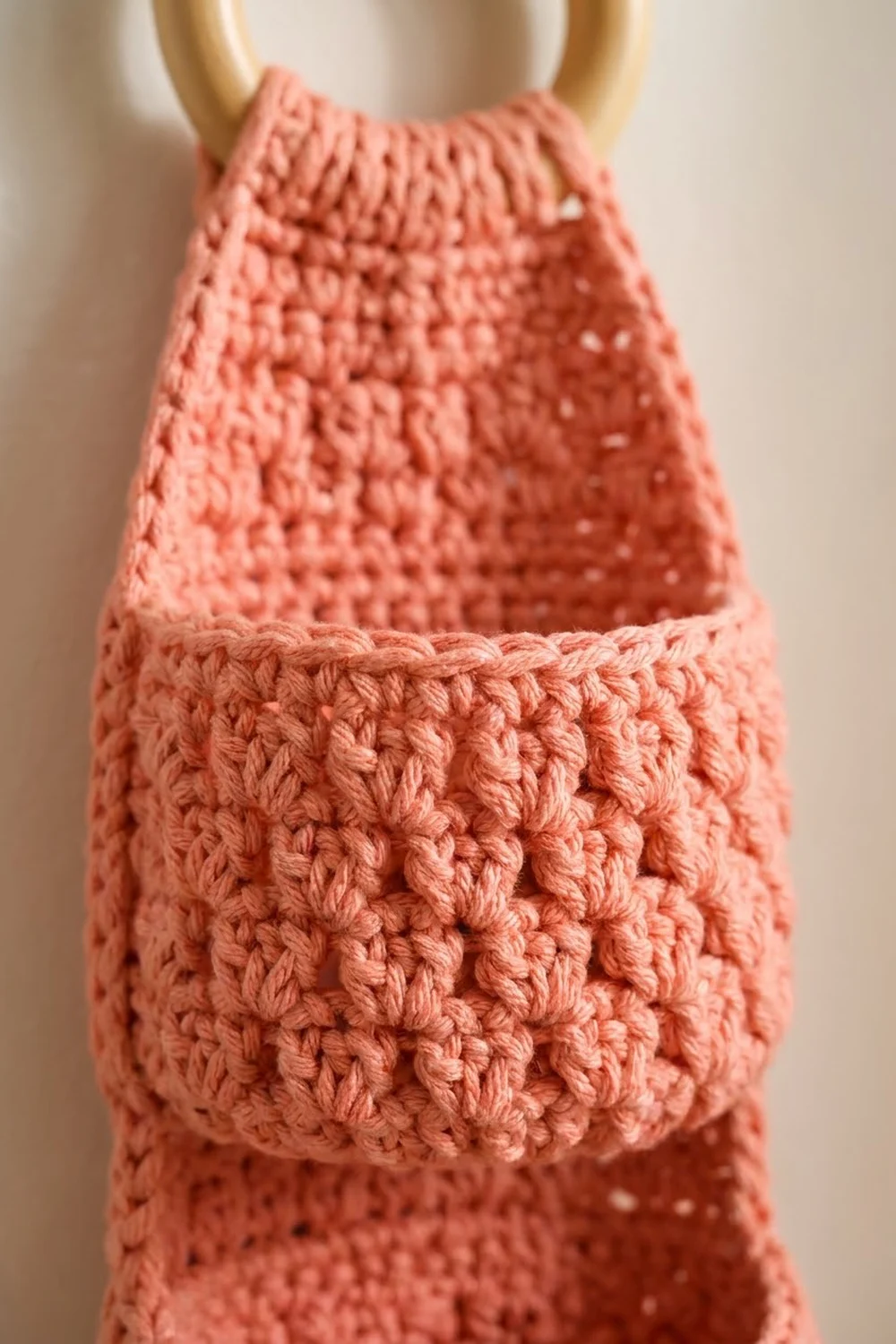 Coral-pink crocheted stuffed toy with textured stitch detailing and tan handles on a neutral background.