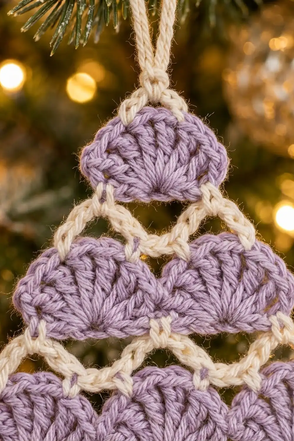 Lavender crocheted ornament with stacked spheres connected by cream rope, hanging against golden holiday bokeh.