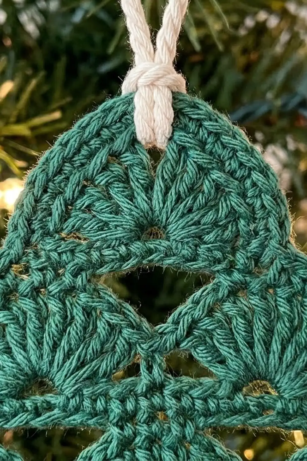 Teal crocheted ornament with textured stitch detail and white rope attachment, photographed against evergreen branches.
