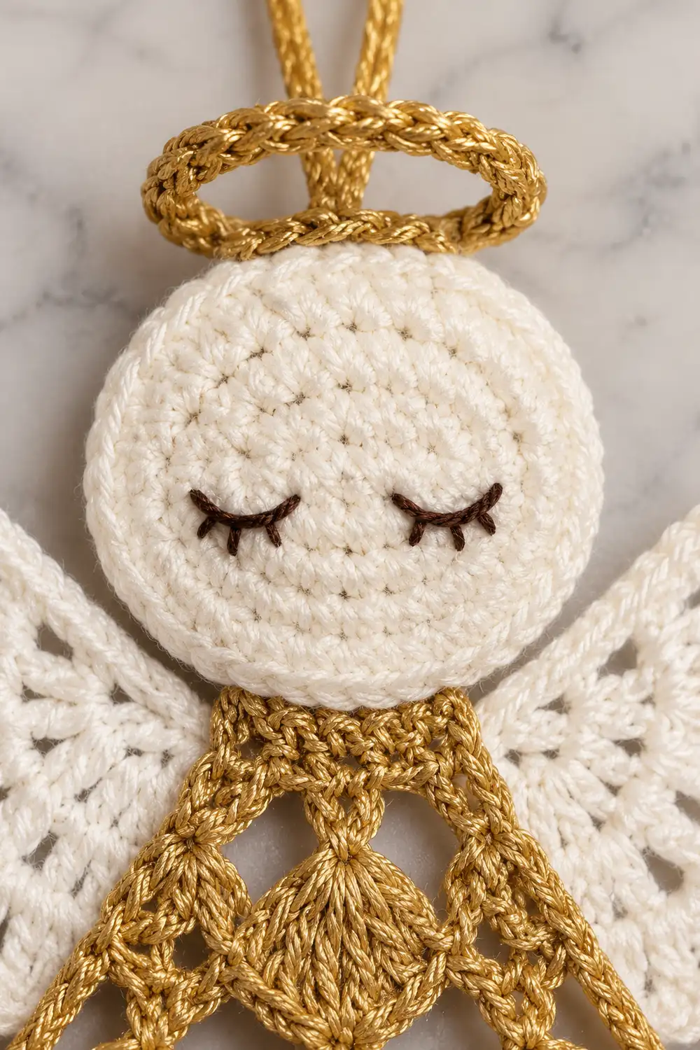 Crocheted white angel ornament with closed eyes, gold yarn accents, and textured wings on marble background.