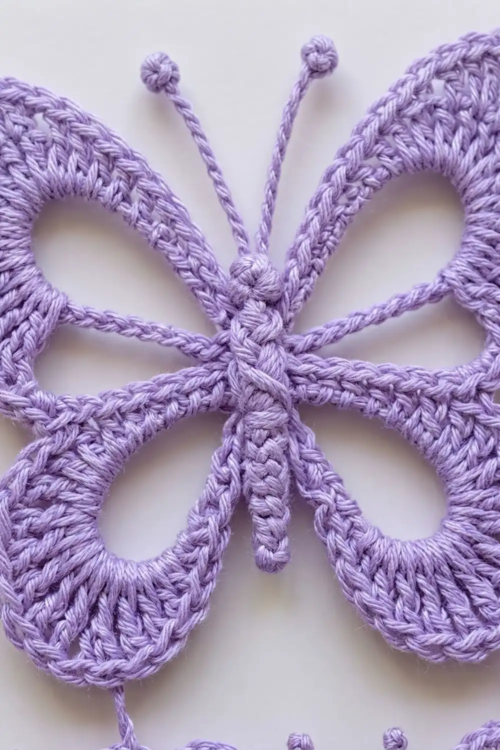 Lavender crocheted butterfly with intricate openwork wings and beaded antennae on a neutral background.