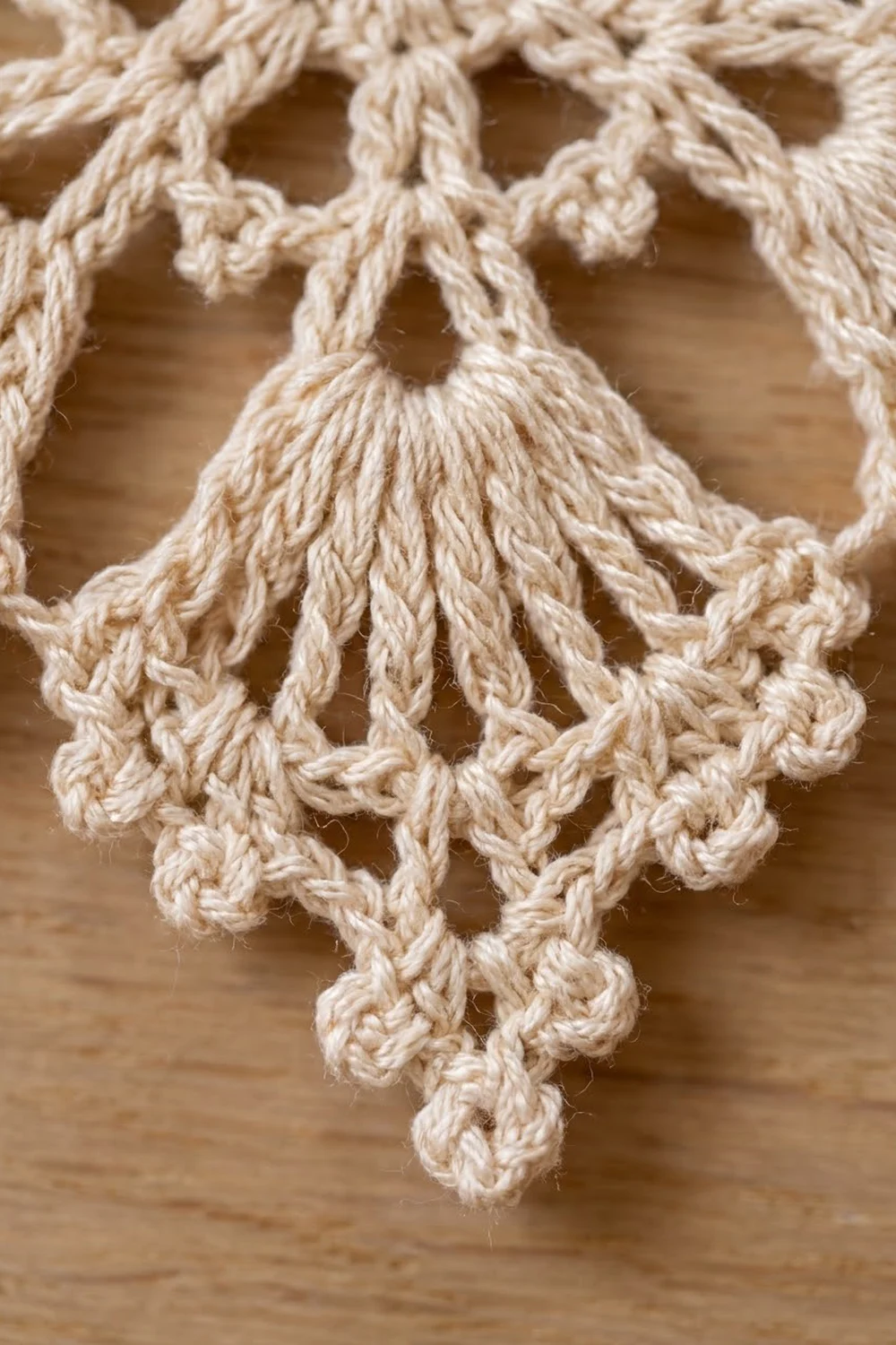 Close-up of cream-colored crocheted lacework with delicate loops and fringe tassels against a blurred tan wooden background.
