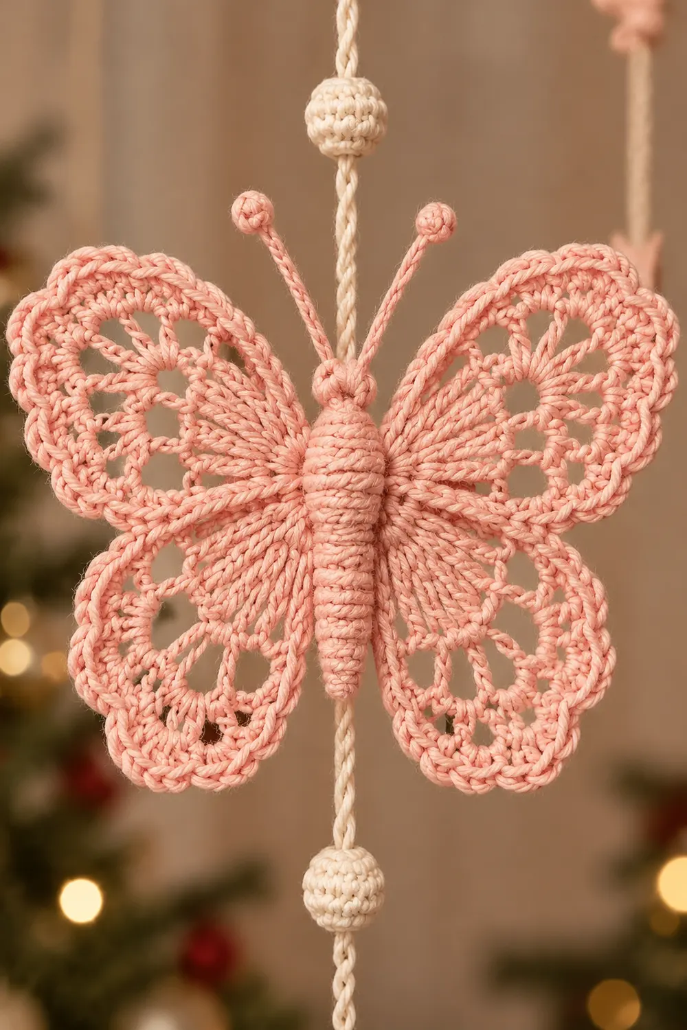 A pink crocheted butterfly ornament with delicate wings hangs from a cream-colored cord with beaded ends.