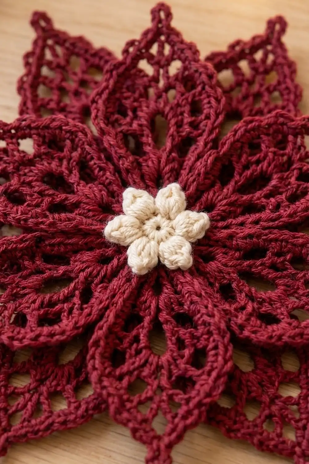 Cream-white six-petaled flower centers a burgundy crocheted bloom with intricate open lace detailing on wooden surface.