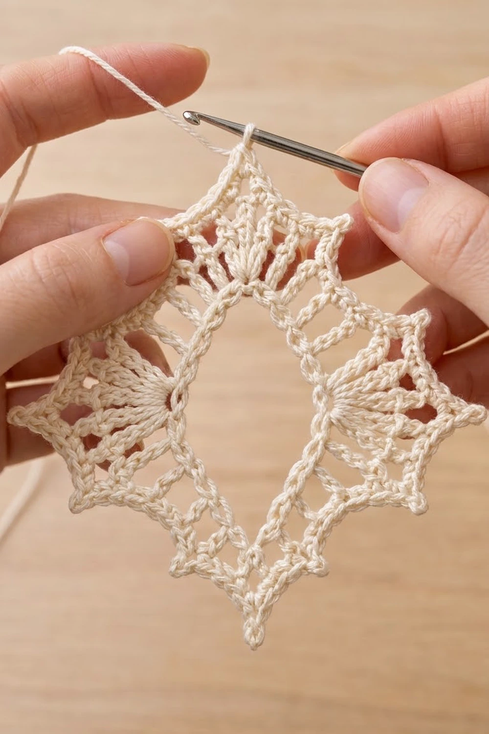 Hands crocheting cream-colored lace with geometric diamond openwork using a metal crochet hook against beige background.