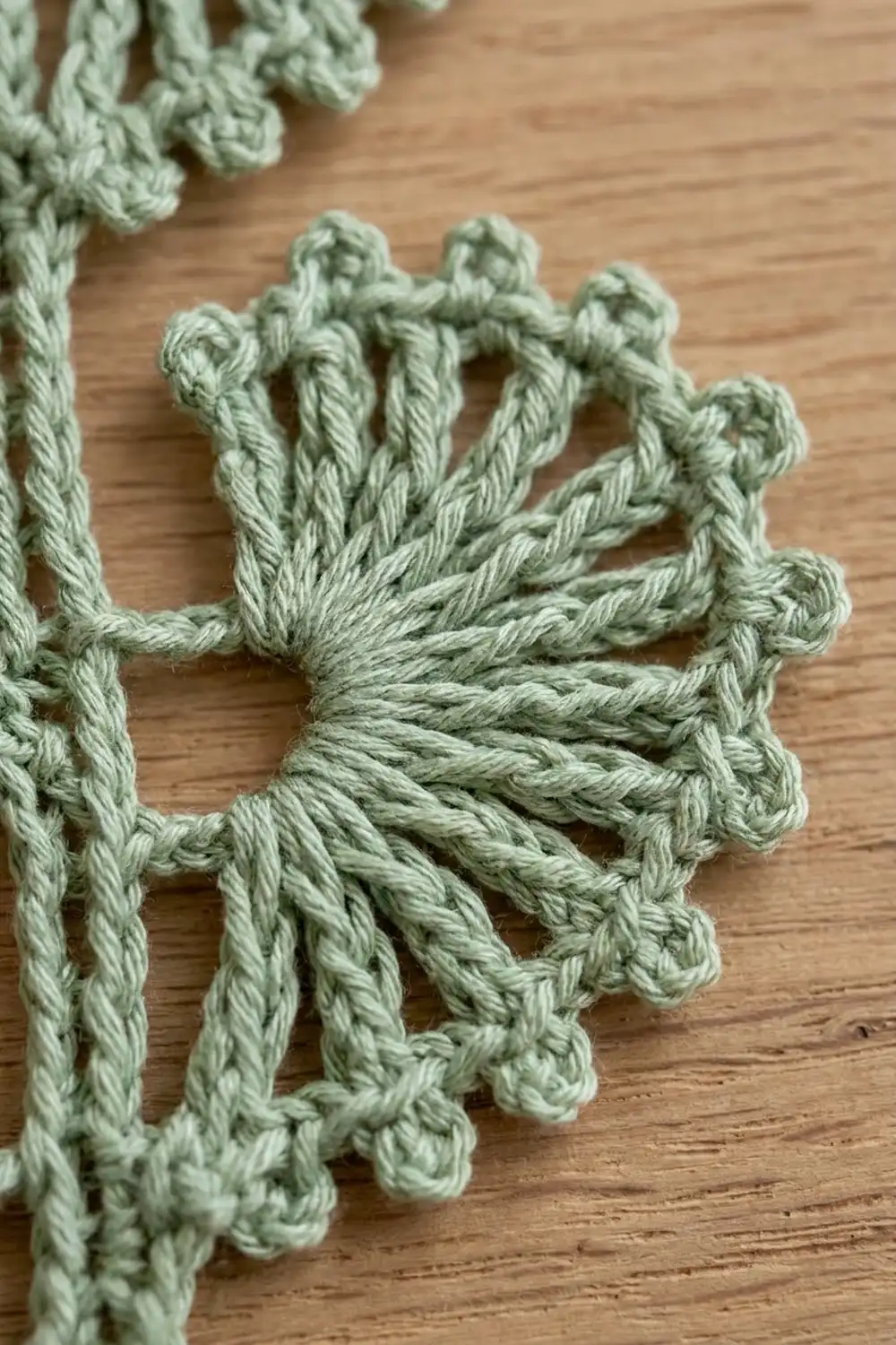 Pale sage-green crochet with detailed knotted stitches and twisted loops arranged on a wooden surface.