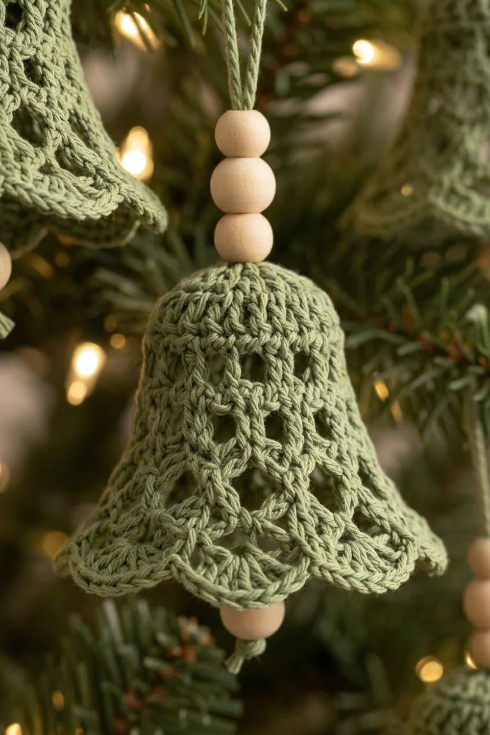 A sage green crocheted bell ornament with wooden beads hanging on an evergreen branch with warm lights.