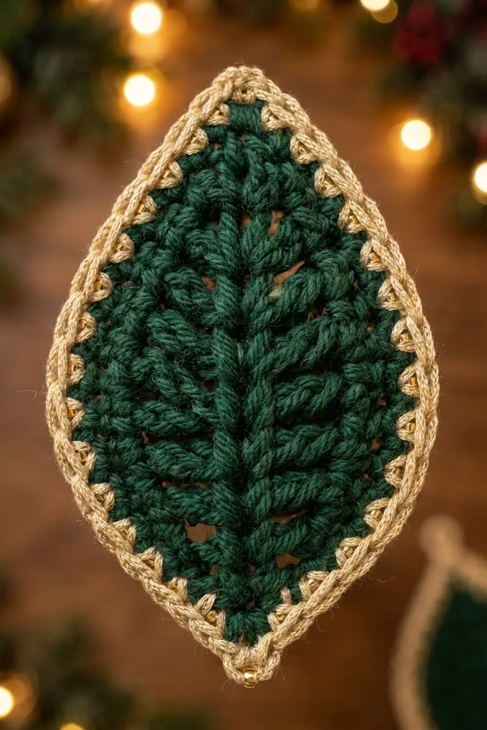 A crocheted ornament with dark green center and tan border hanging against warm holiday lights.