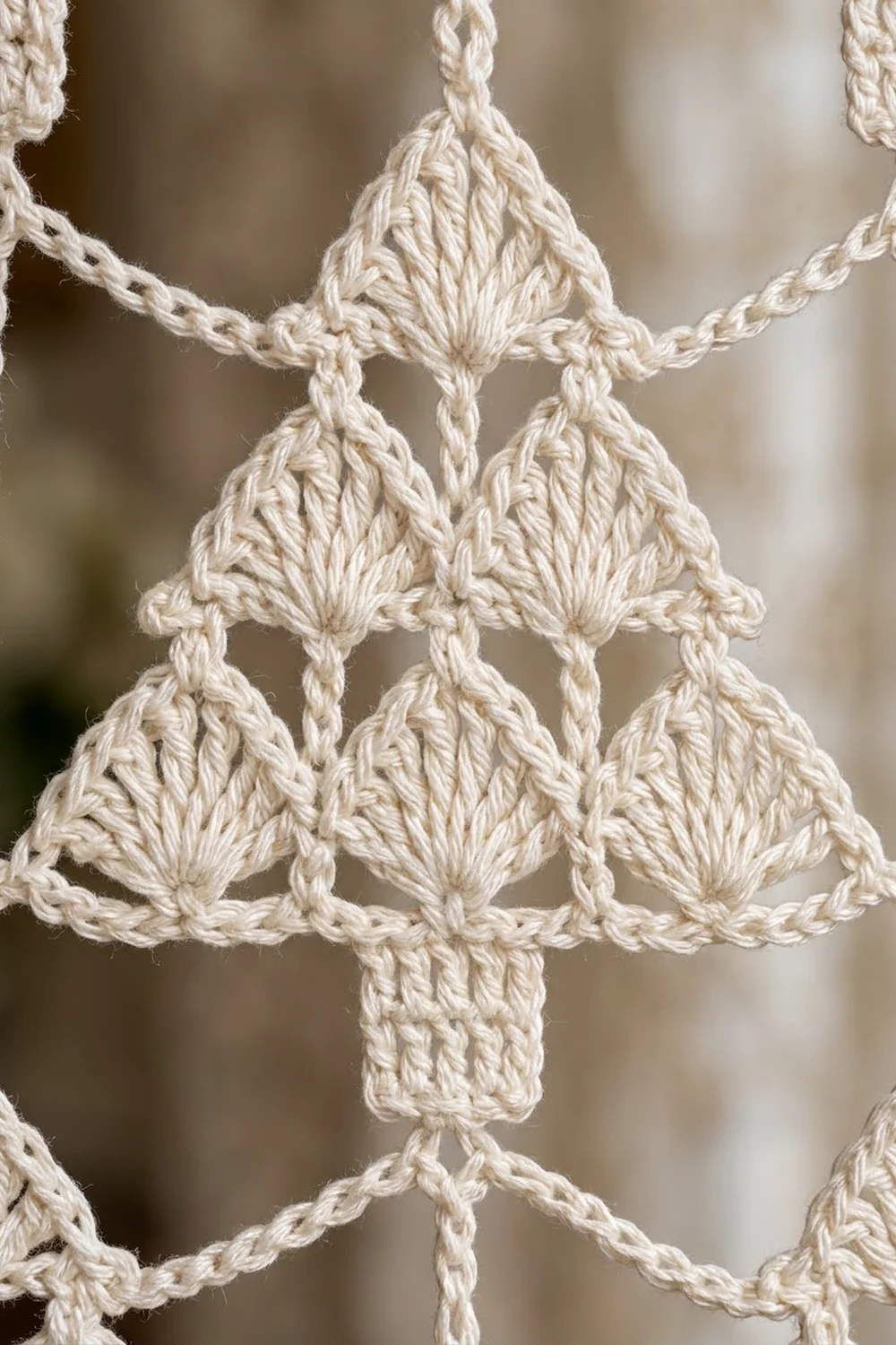 Close-up of ivory crochet lace with symmetrical triangle motifs and rope stitching on a blurred background.