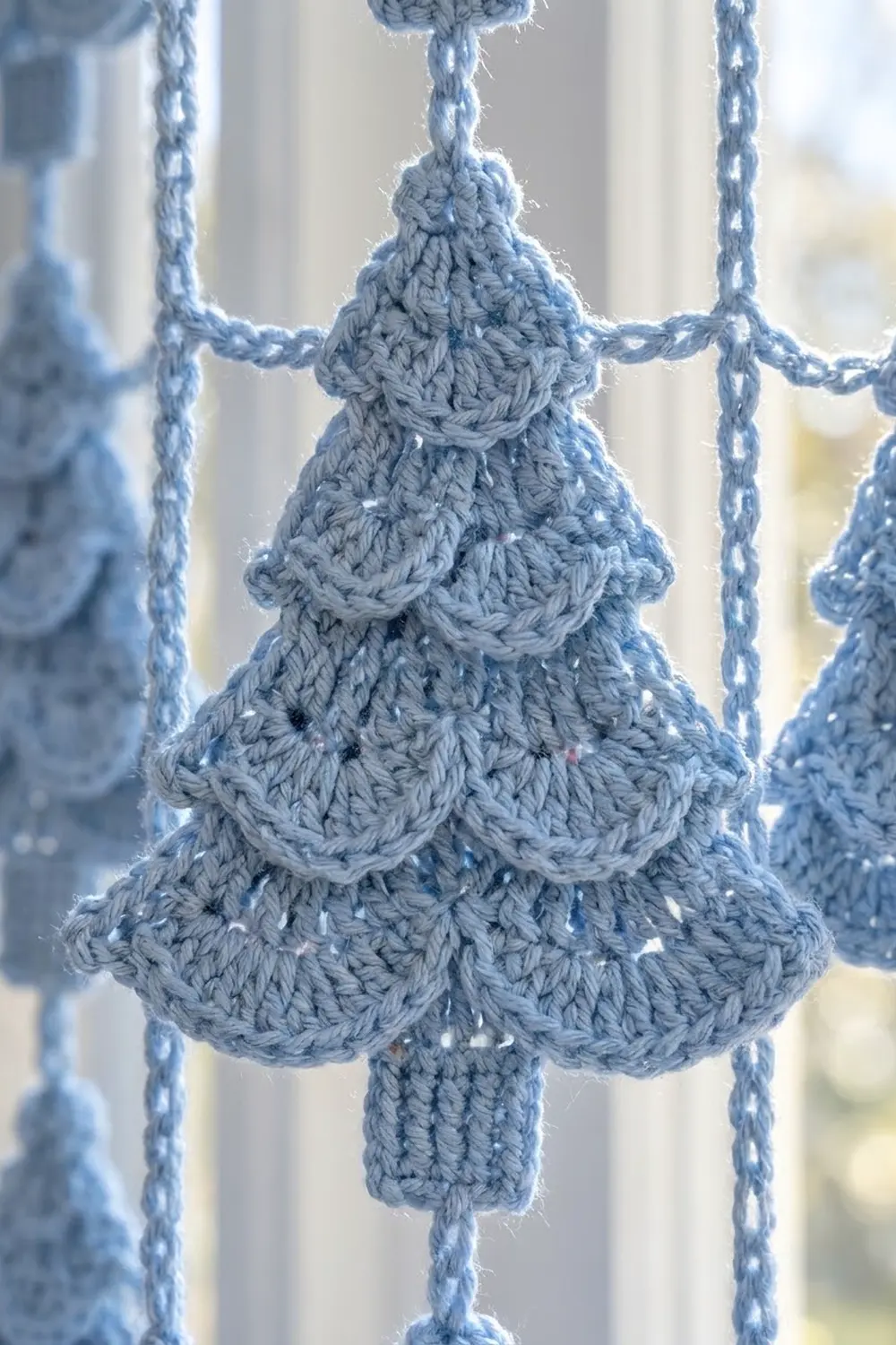 Hand-crocheted gray Christmas tree ornament with ruffled layers hanging from blue twine against a bright window.