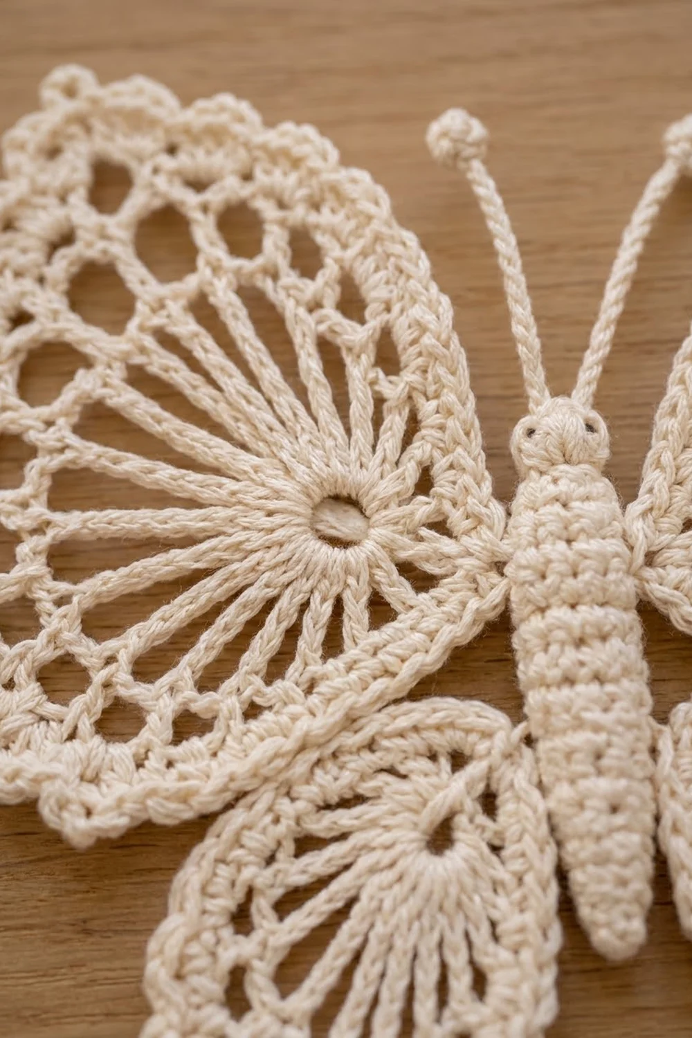 Cream-colored crochet butterfly featuring intricate lacework wings and textured body with thin antennae on wood.