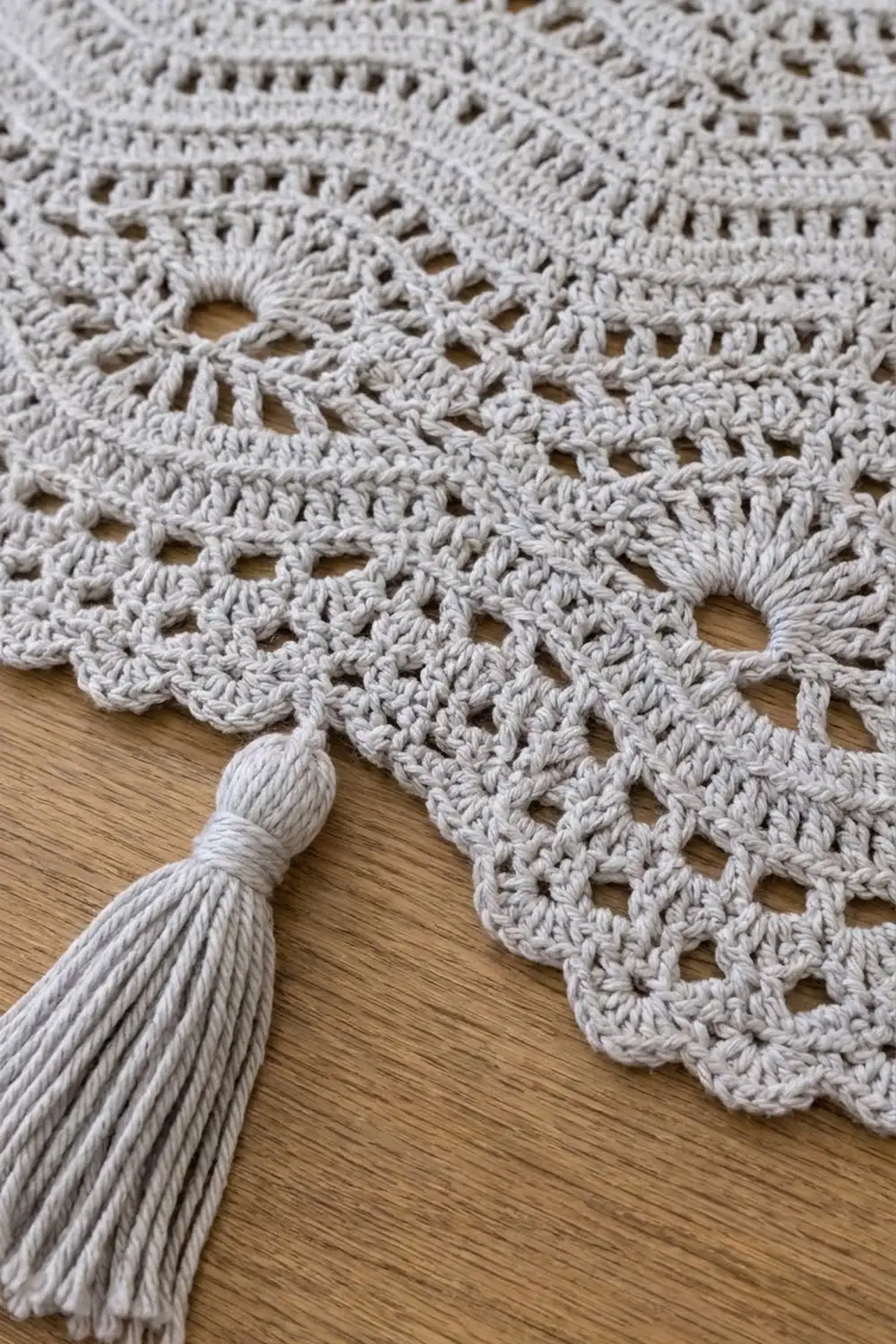 Cream-colored crocheted piece with delicate openwork lace pattern and decorative tassel on wooden background.