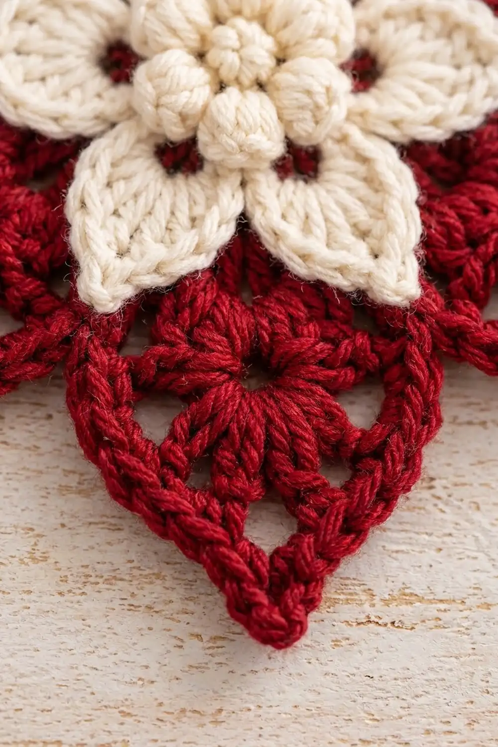 Cream-colored crochet flower with petals above a deep-red crocheted heart on a weathered beige surface.