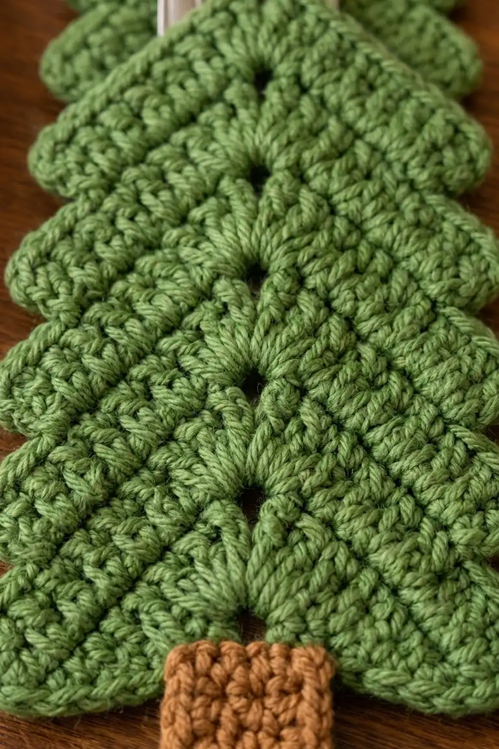 Sage-green crocheted Christmas tree displaying textured popcorn stitches with brown trunk on wooden surface.