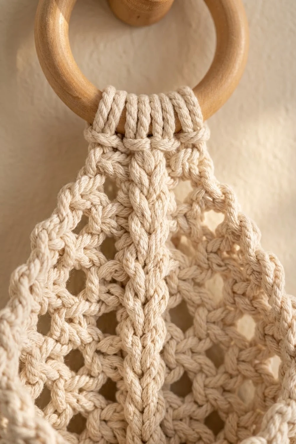 Cream macramé hanging with intricate rope knotwork and natural wood ring on neutral beige background.