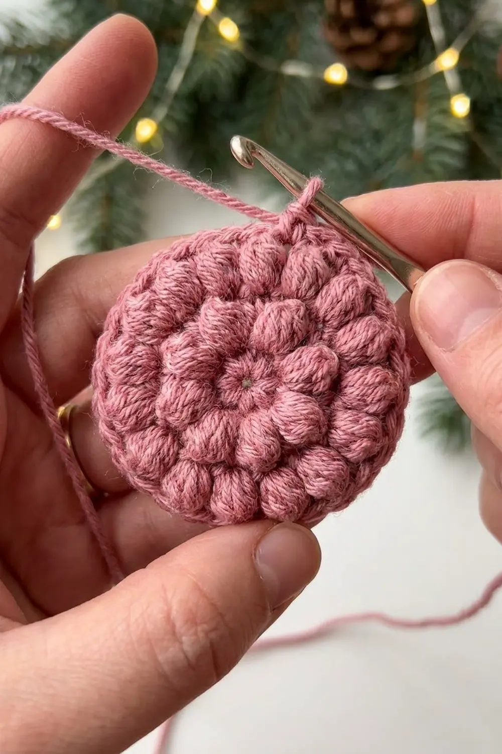 Close-up of hands holding a pink crocheted ball ornament with a crochet hook against a festive evergreen background.