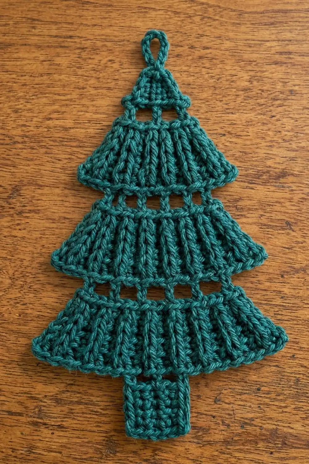Handmade teal crochet Christmas tree ornament with three layered sections and hanging loop on wooden surface.