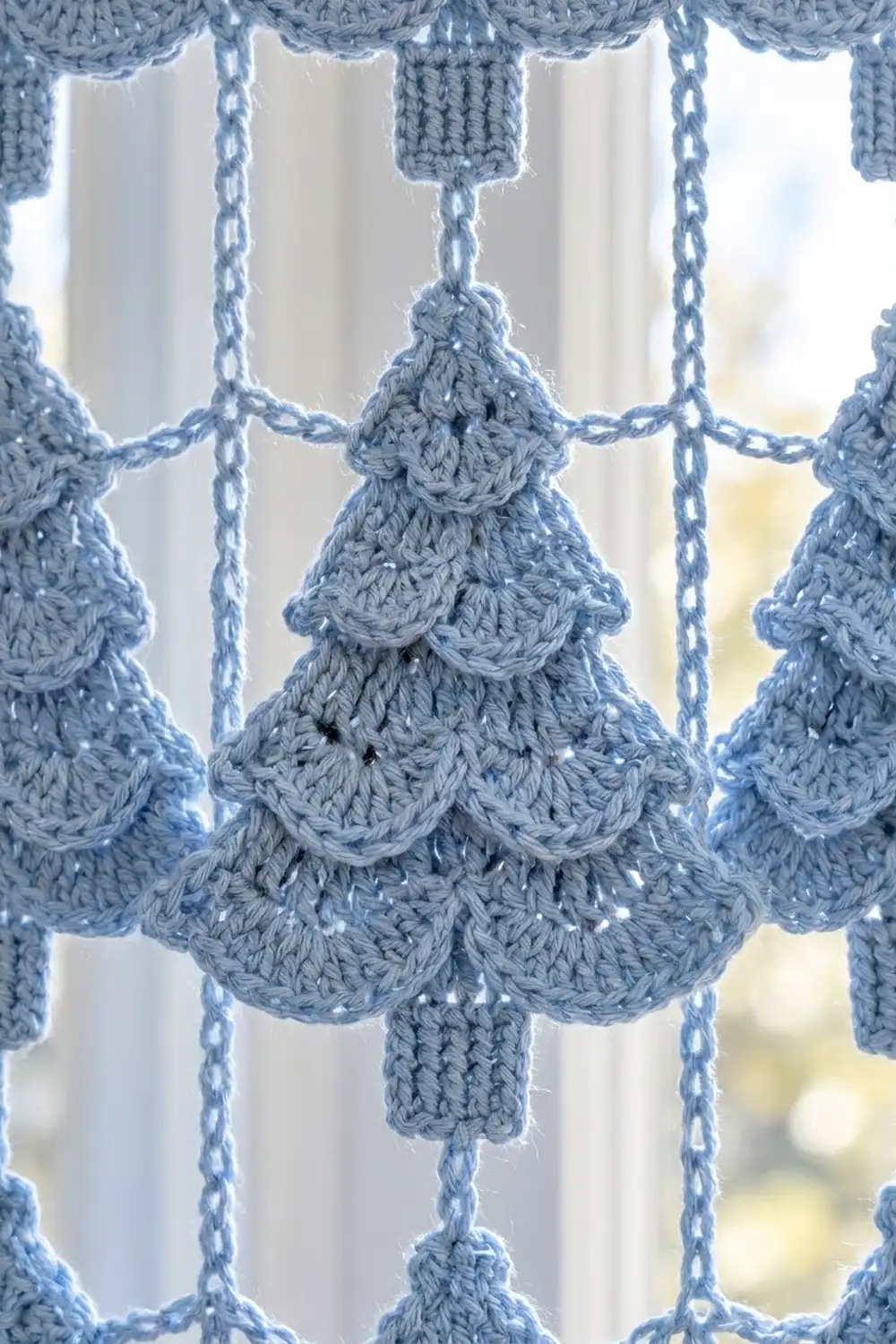 Dusty blue crocheted Christmas tree ornament with textured stitching, hanging from chains against a bright window backdrop.