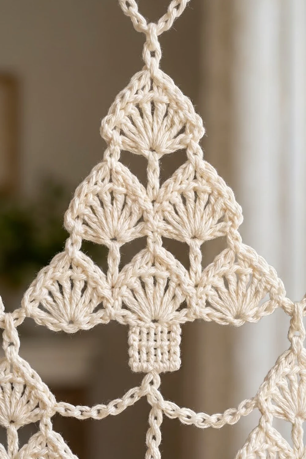 Cream-colored crocheted Christmas tree ornament with shell stitch detailing, hanging from a chain against a blurred interior background.