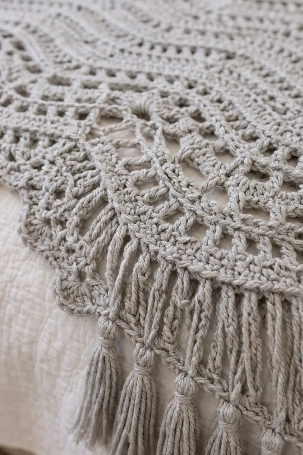 Close-up of cream-colored crocheted lace fabric showing open stitching pattern and knotted fringe hem.