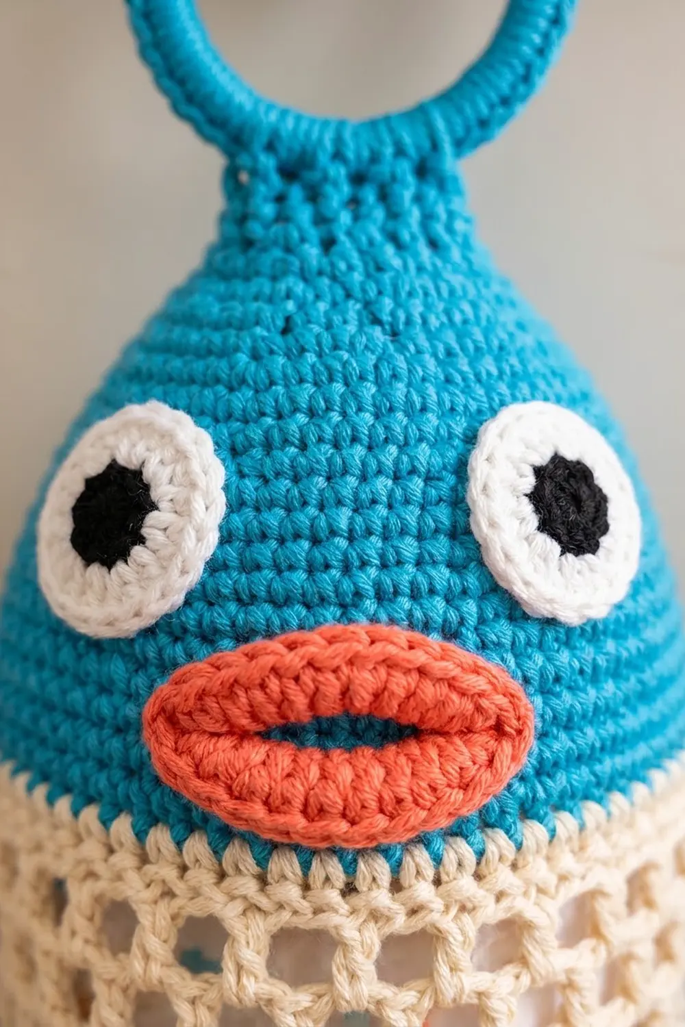 Turquoise crocheted creature with white-and-black eyes, coral-red stitched smile, and cream-colored bottom.