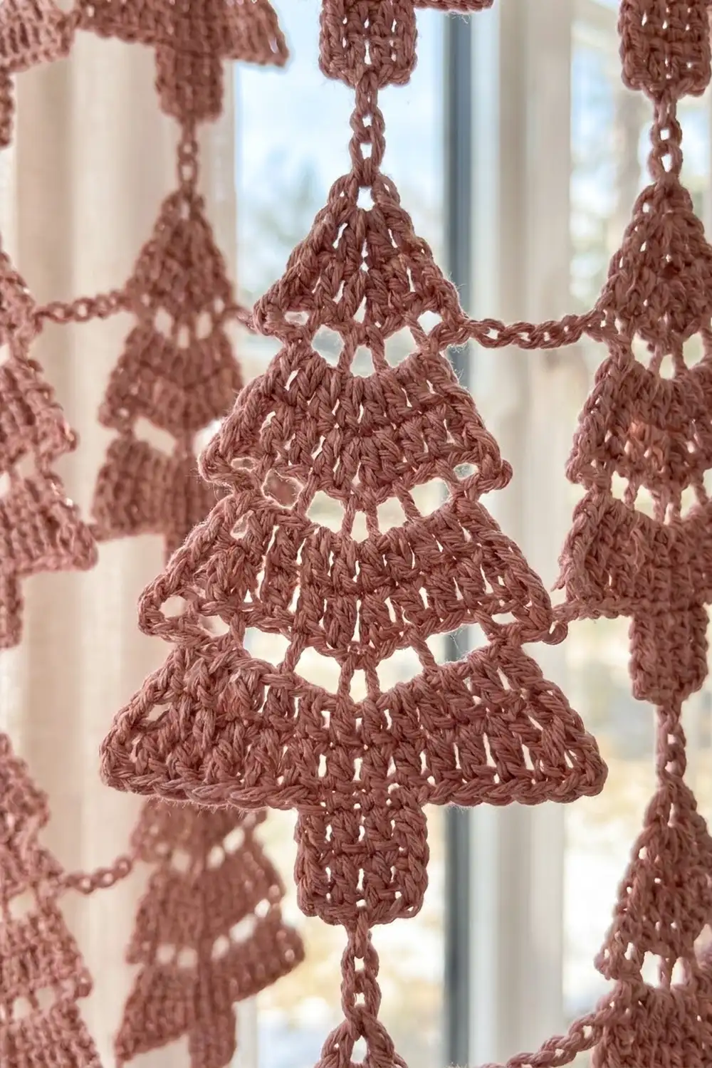 Dusty rose crocheted tree ornaments with lace-like openwork details hang backlit in a window.